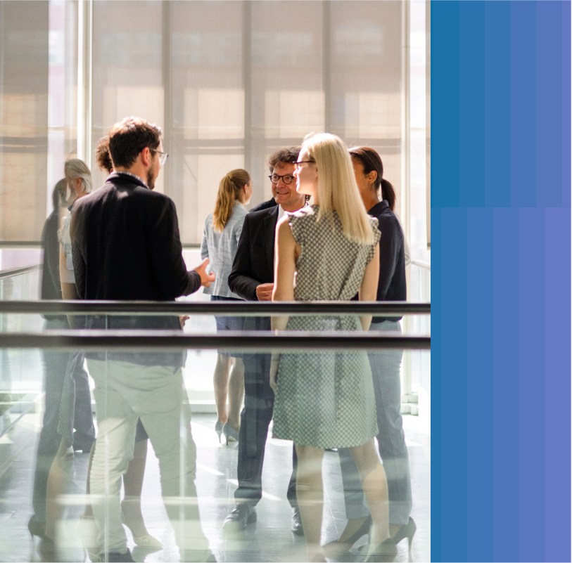 photo, employees chat at a gathering in a large office hallway. They are standing near waist-high glass guard rails with brown banisters. A large building can be seen through the open window nearby.