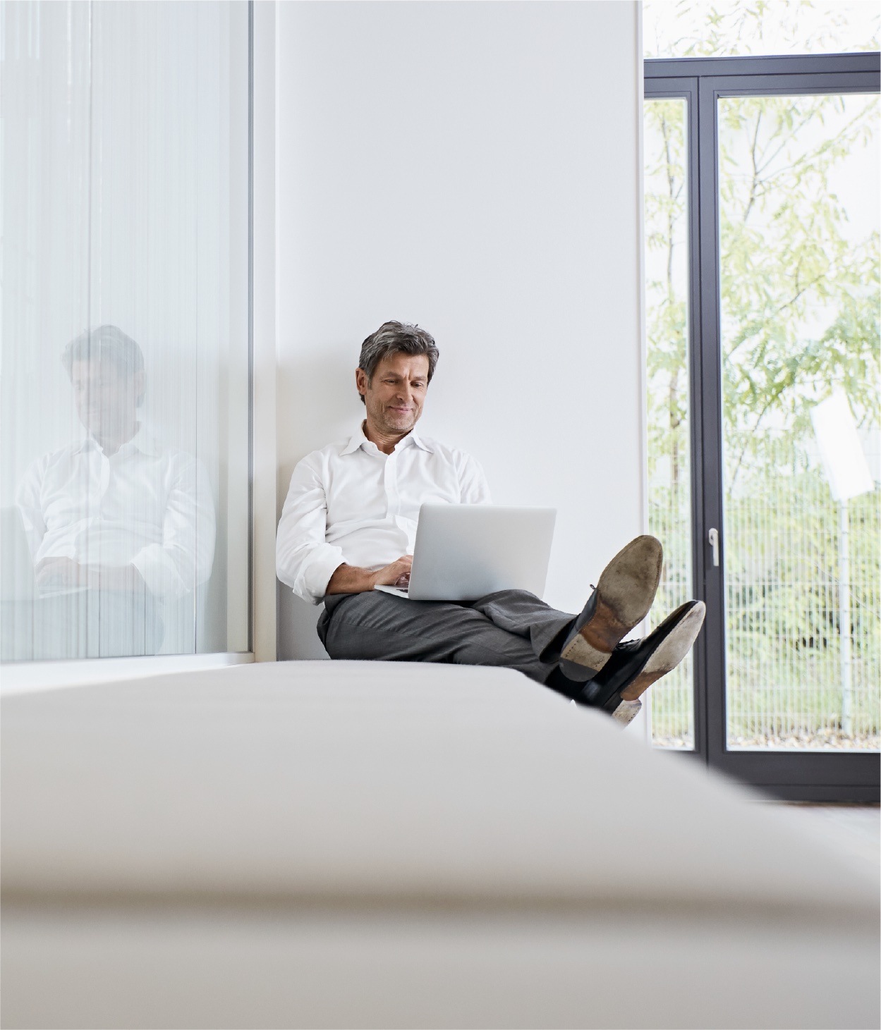 Man works on his laptop while reclining on a bench inside an office hallway.